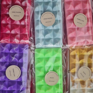 Puede incluir: Una variedad de ceras perfumadas de colores en envases transparentes. Los colores incluyen rojo, azul, rosa, morado, verde y amarillo. Cada cera tiene una superficie texturizada y una etiqueta redonda con texto. Las etiquetas dicen "LES FONDANTS D'ALIYAH" y otra información.