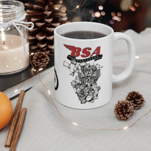 Könnte beinhalten: Eine weiße Keramiktasse mit einer Schwarzweißillustration eines Motorradmotors und dem Text "BSA Motorcycles" in roten Buchstaben. Die Tasse ist mit Kaffee gefüllt.