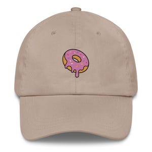 Op de afbeelding: Beige baseballpet met een geborduurd roze donutontwerp. De donut heeft roze glazuur met hagelslag en een gele omtrek. De pet heeft een gebogen klep en een verstelbare band.