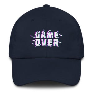 May include: A navy blue baseball cap featuring the text "GAME OVER" in white, pixelated font with a glitch effect. The cap has a curved peak and a classic six-panel construction. A stylish accessory.