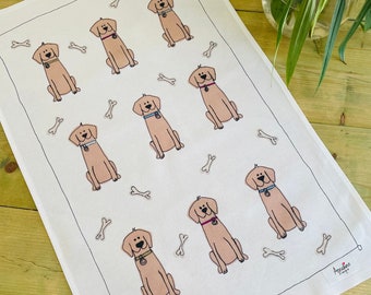 May include: White tea towel with a repeating pattern of brown Labrador Retriever dogs and white bones. The tea towel has a small white tag with the text "Apples & Things" in black.