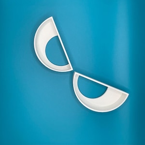 May include: Two white, semi-circular wall shelves with a rounded interior, set against a teal background. The shelves are made of a smooth, matte material and are positioned at an angle.