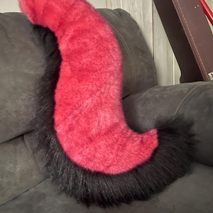 May include: A long, curved faux fur tail featuring a vibrant magenta-pink center section that transitions into deep black fur at both the top and bottom ends. The plush accessory has a thick, shaggy texture and a distinct S-shaped silhouette.