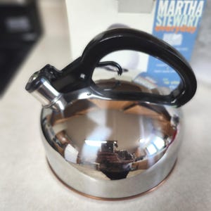 May include: A polished stainless steel tea kettle with a black handle and a whistle spout. The kettle features a copper-colored base and is positioned in front of a white product box with the text Martha Stewart Everyday printed in blue and orange.