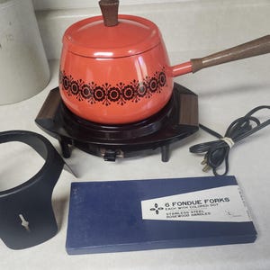 May include: An orange fondue pot with a brown floral pattern, a wooden handle, and a matching lid. It sits on a dark brown electric heating base with a power cord. Included is a black metal stand and a blue box labeled 6 FONDUE FORKS.
