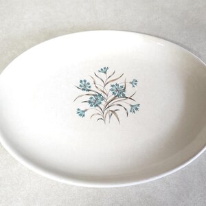 May include: An oval, off-white serving platter with a floral design. The center features a cluster of blue flowers with brown stems and leaves. The rim has a subtle, textured edge, adding a vintage feel to the dish.