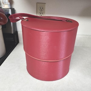 May include: A vintage red hat box with a matching handle. The cylindrical box is made of a textured material and has a lid. The box is divided into two sections with a thin red band separating them. The handle is attached with gold-colored hardware.