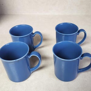 May include: Four blue ceramic mugs with curved handles. The mugs have a simple, classic design and a glossy finish. The mugs are arranged on a light-colored surface, perfect for enjoying coffee, tea, or other beverages.