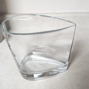 May include: A clear, modern glass vase with a unique, asymmetrical design. The vase has a curved top and a gently swirling base, creating an elegant and contemporary look. The clear glass allows for visibility of the contents.