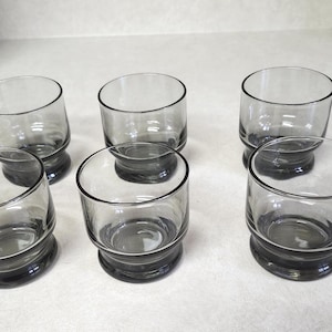 May include: Six gray-tinted drinking glasses. Each glass has a rounded, cylindrical shape with a thick, dark gray base. The glasses are arranged on a light-colored surface, showcasing their simple, yet elegant design. Ideal for serving beverages.