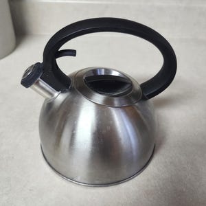May include: A stainless steel whistling tea kettle with a rounded body, a black arched handle, and a black spout cap. The kettle features a circular lid with a black handle in the center and a metallic finish, designed for stovetop water heating.