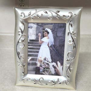 May include: A silver-colored picture frame with a decorative floral design. The frame holds a photograph of a bride in a white dress and veil. The text "ORAL INNOCENCE" and "LENOX" are visible.