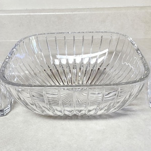 May include: A clear glass serving bowl with a square shape and vertical ribbed design, flanked by two smaller matching square glass dishes. The glassware is set on a light-colored countertop.