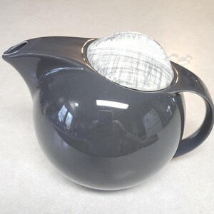 May include: A glossy black spherical teapot featuring a curved spout and a handle integrated into the body. The top of the teapot is covered by a circular lid with a white surface marked by fine, intersecting black lines in a crosshatch pattern.