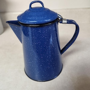 May include: A deep blue enamel coffee pot featuring a white speckled pattern throughout the exterior. The pot includes a matching lid with a small handle, a side spout, and a curved metal handle attached with a silver-colored metal hinge.