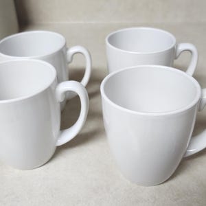 May include: Four white ceramic mugs with curved handles are arranged on a light-colored surface. The mugs have a simple, rounded design, suitable for coffee or tea. The mugs are evenly spaced, showcasing their clean, minimalist aesthetic.