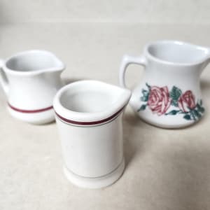 May include: Three white ceramic cream pitchers. Two have a red stripe around the base, and one has a floral rose design. The pitchers are small and have a classic design.