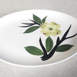 May include: An oval white ceramic platter featuring a hand-painted design of a pale yellow flower with dark brown centers, surrounded by dark green leaves and black branches extending across the surface of the dish in a simple, clean composition.