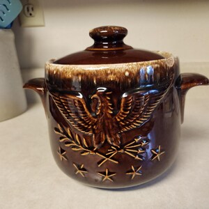 May include: A brown and white glazed ceramic crock pot with a lid. The crock pot features a brown eagle with outstretched wings, a branch with leaves, and stars.