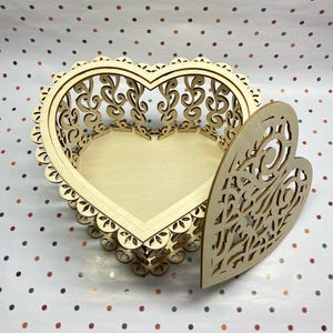 May include: A wooden heart-shaped box with a delicate, intricate design. The box is open, revealing the empty interior. The box is made of light-colored wood and has a natural finish.