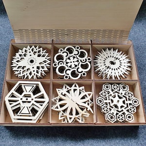 May include: A wooden box with six laser-cut wooden ornaments. The ornaments are in the shapes of snowflakes, stars, and geometric designs. The box is made of light wood and has a natural finish.