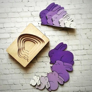 Può includere: Un puzzle in legno con un set di forme di conigli viola e bianchi. I conigli sono di diverse dimensioni e si incastrano l'uno nell'altro. Il puzzle è in legno e ha una finitura naturale.