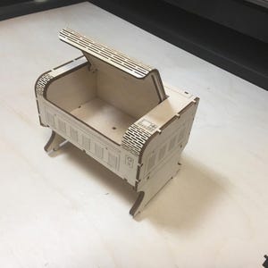Op de afbeelding: Een houten doos met een scharnierend deksel, de doos is versierd met een ontwerp dat lijkt op een trein. Het deksel is open en de binnenkant van de doos is leeg.