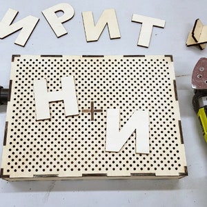 Könnte beinhalten: Eine perforierte Holzkiste mit den Buchstaben "H" und "И" darauf. Zusätzliche Holzbuchstaben und ein Elektrowerkzeug mit Schleifpad sind ebenfalls vorhanden. Die Gegenstände befinden sich auf einer hellgrauen Oberfläche.