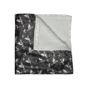 May include: A black and white patterned blanket featuring a bat and swirl design. The blanket has a soft, plush texture and a white, fluffy interior. The blanket is folded over, revealing the white interior.