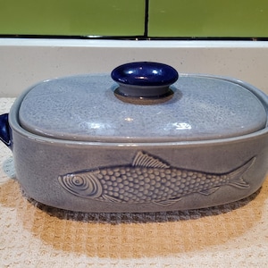 Könnte beinhalten: Hellblaue Keramik-Auflaufform mit Deckel. Die Schüssel hat ein Fischdesign an der Seite und einen dunkelblauen Knopf auf dem Deckel. Die Schüssel ist etwa 25 cm lang.