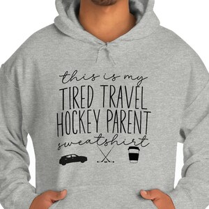 Op de afbeelding: Grijs hoodie met de tekst "This is my tired travel hockey parent sweatshirt" en een afbeelding van een auto, hockeysticks en een koffiebeker.