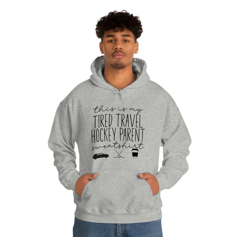 Op de afbeelding: Grijs hoodie met de tekst "This is my tired travel hockey parent sweatshirt" en een afbeelding van een auto, hockeysticks en een koffiebeker.