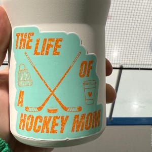 May include: A white tumbler with a teal sticker that reads "THE LIFE OF A HOCKEY MOM" in orange. The sticker features hockey sticks, a beanie, a coffee cup, and a hockey puck. The Hockey Mom Shop logo is visible.