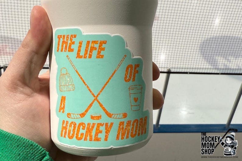 May include: A white tumbler with a teal sticker that reads "THE LIFE OF A HOCKEY MOM" in orange. The sticker features hockey sticks, a beanie, a coffee cup, and a hockey puck. The Hockey Mom Shop logo is visible.