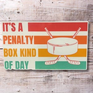 May include: A retro-style graphic with a hockey puck and crossed hockey sticks. The text reads "It's a Penalty Box Kind of Day".