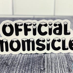 May include: Black and white sticker with the text "Official Momsicle" in a stylized font with white icicles on top of each letter.