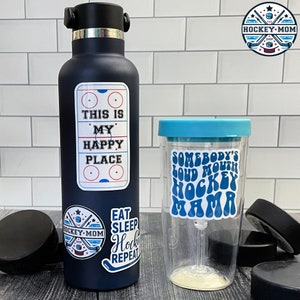 May include: A navy blue water bottle and a clear tumbler with hockey-themed stickers. The water bottle has stickers that say "This is my happy place" and "Eat Sleep Hockey Repeat." The tumbler says "Somebody's Loud Mouth Hockey Mama."