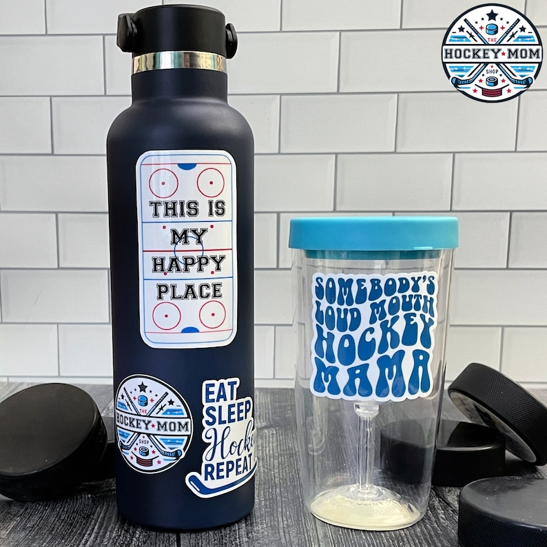 May include: A navy blue water bottle and a clear tumbler with hockey-themed stickers. The water bottle has stickers that say "This is my happy place" and "Eat Sleep Hockey Repeat." The tumbler says "Somebody's Loud Mouth Hockey Mama."