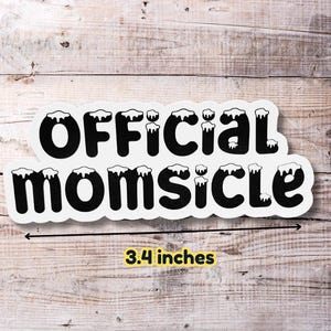 May include: White sticker with black text that reads "OFFICIAL MOMSICLE" with a white outline and a dripping effect. The sticker is 3.4 inches wide and is on a wood background.