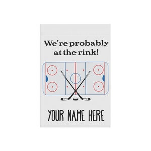 K&ouml;nnte beinhalten: Eine wei&szlig;e Gartenflagge mit einem Eishockeyfeld und zwei gekreuzten Schl&auml;gern. Der Text "We're probably at the rink!" befindet sich &uuml;ber dem Feld und "YOUR NAME HERE" darunter.
