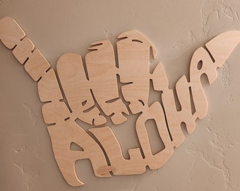May include: A wooden sign in the shape of a hand making the shaka sign. The sign is carved with the word "Aloha" in a stylised font.