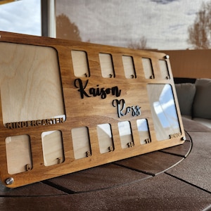 May include: A wooden photo display with 11 picture frames, a large frame at the top, and smaller frames below. The display is labeled "Kindergarten" and has the name "Kaisen Ross" written on it.