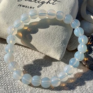 May include: A bracelet made of opalescent, round beads. The beads are a soft white color with subtle blue and white striations. The bracelet is resting on a textured surface next to a small bag with the text "Sealight JEWELRY".