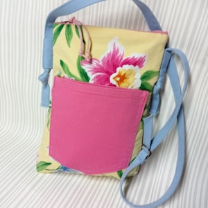 Può includere: Una borsa a tracolla gialla con motivo floreale, una tasca rosa e una tracolla blu chiaro. La borsa ha una chiusura a zip e un piccolo anello in cima.