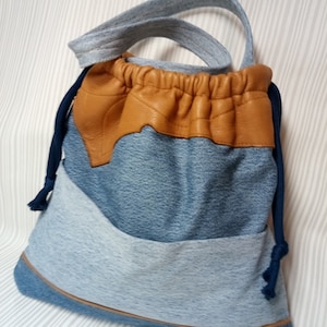 Puede incluir: Una bolsa de mano de lona azul con detalles de cuero marrón. La bolsa tiene un cierre de cordón y una sola correa para el hombro.