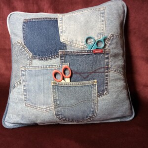 May include: Square denim pillow made from repurposed jeans. The pillow features various denim patches and pockets. A pair of small blue scissors and a pair of red scissors are placed in the pockets. The pillow is on a burgundy surface.