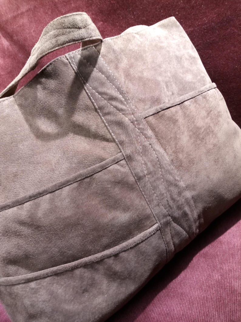 May include: A large, gray suede tote bag with a stitched design. The bag has a wide, flat handle and multiple compartments.