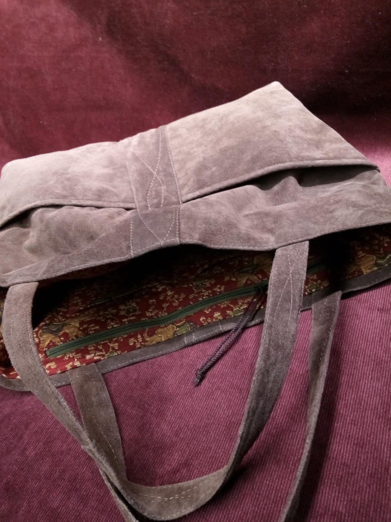 May include: A large, brown suede tote bag with a zippered pocket and a patterned fabric lining. The bag has two long straps.
