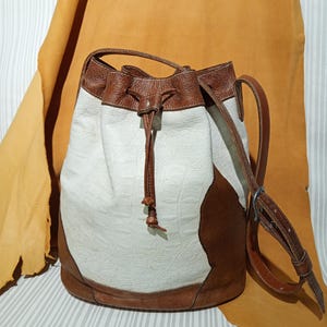 May include: A bucket bag featuring a white, textured, crocodile-embossed leather body contrasted with smooth brown leather panels at the base and top. The bag includes a brown leather drawstring closure and an adjustable brown leather shoulder strap.