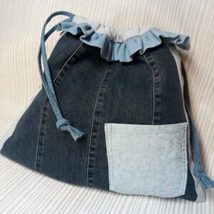 Puede incluir: Una bolsa de mezclilla con cordón, con un bolsillo floral azul claro y una parte superior con volantes. La bolsa está hecha de mezclilla reciclada y tiene un cierre de cordón. Un accesorio único y ecológico.